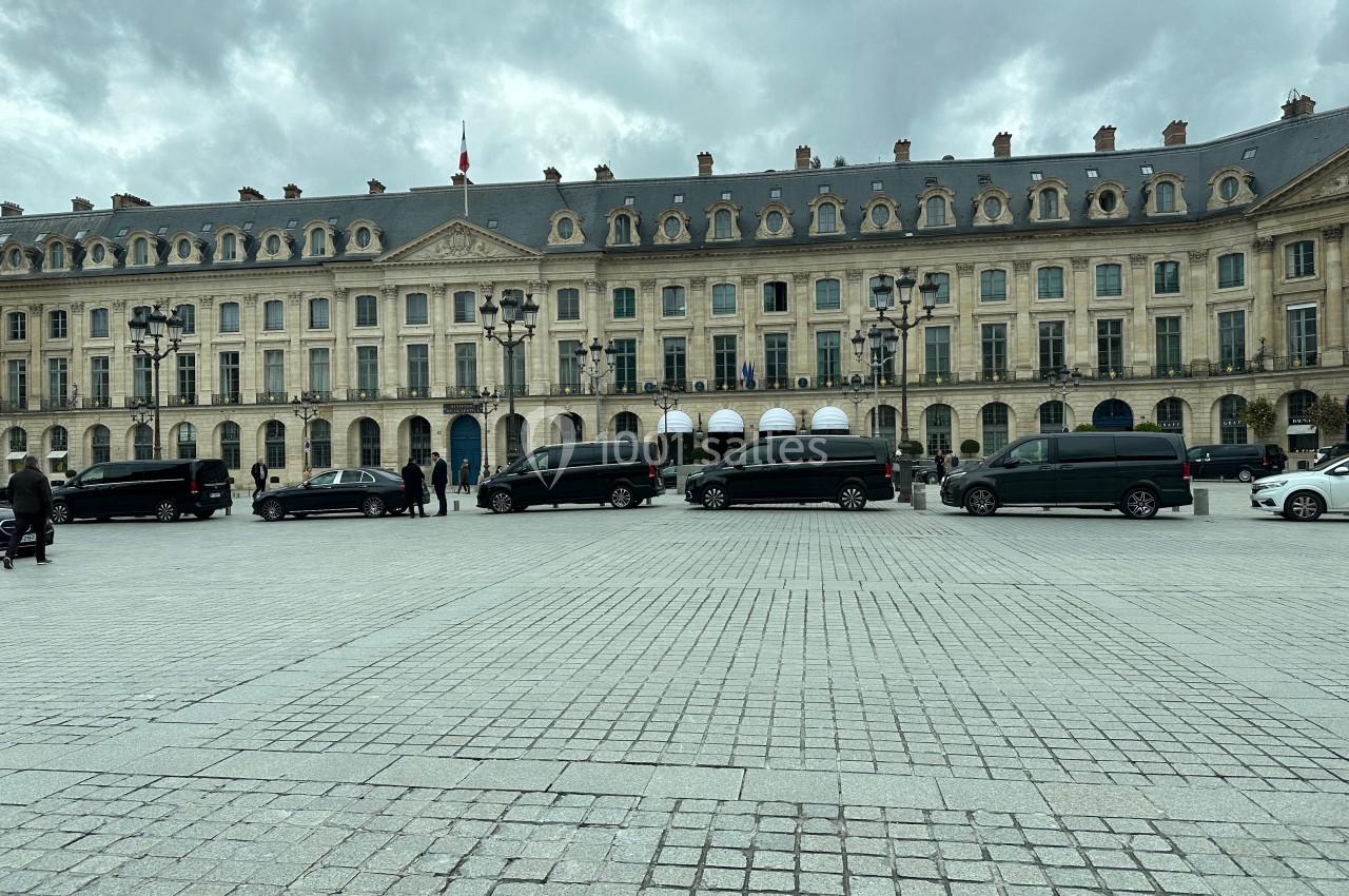 Façade d'un bâtiment historique avec des voitures noires alignées sur une place pavée sous un ciel nuageux.