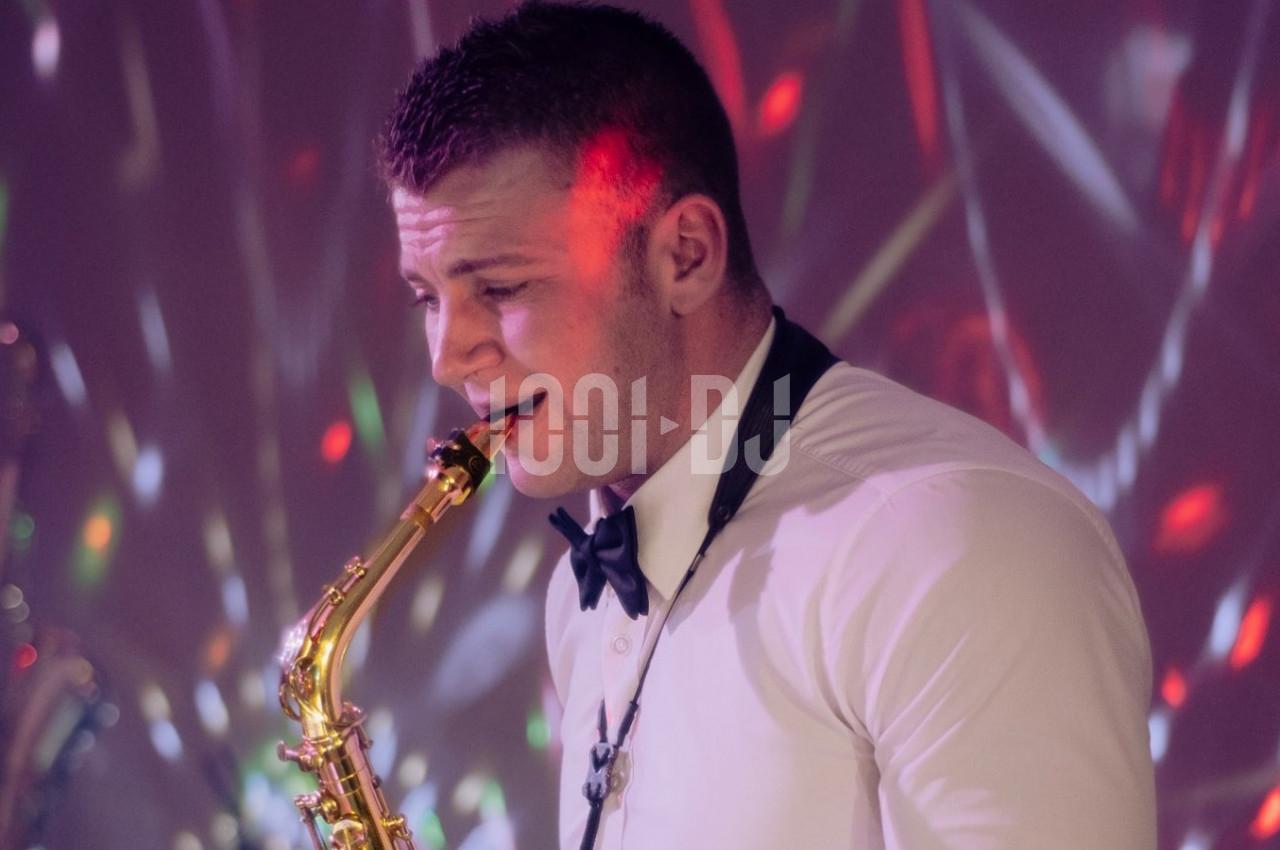 Un musicien en chemise blanche et nœud papillon joue du saxophone sous des lumières colorées.