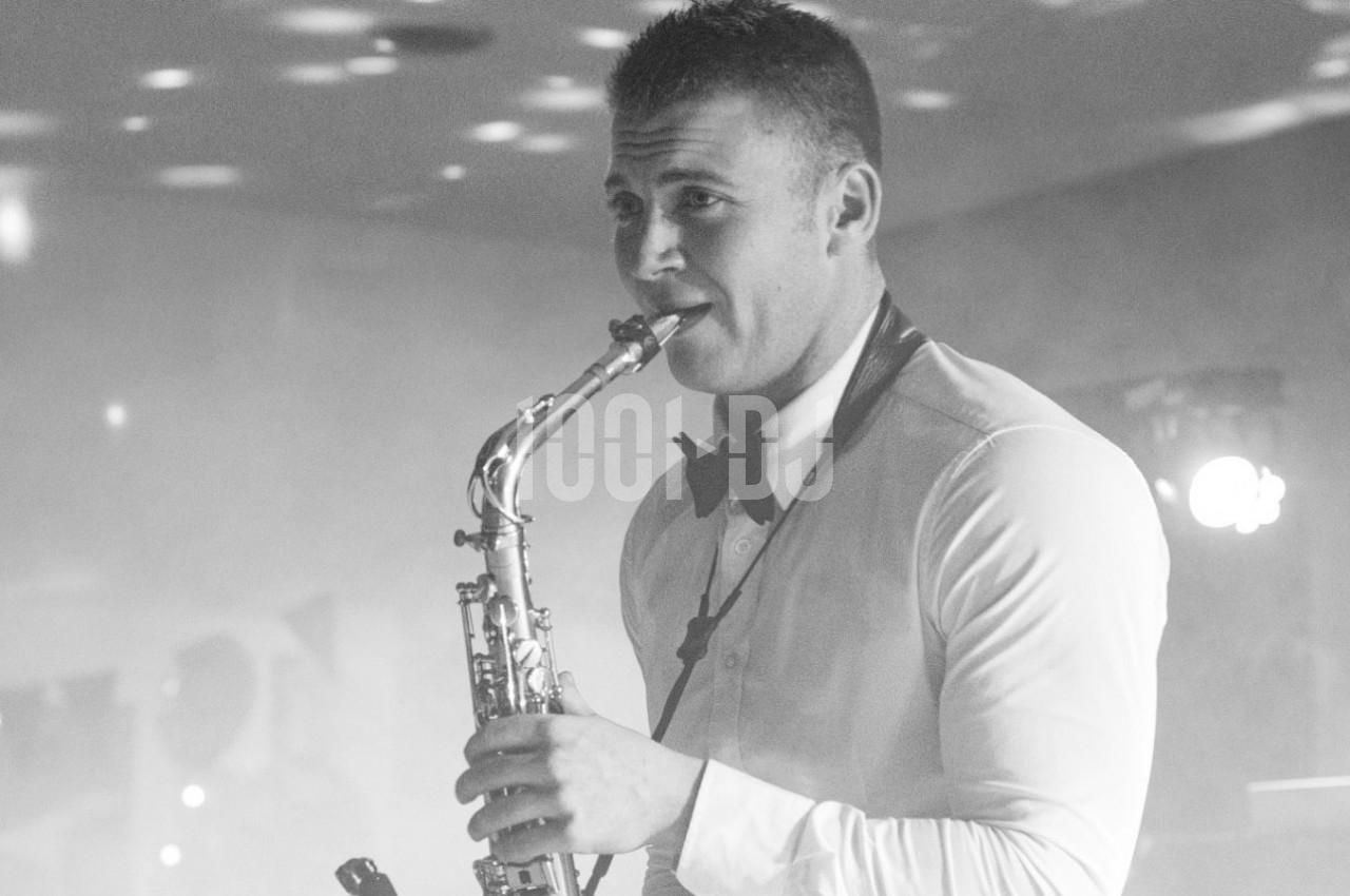 Un musicien en chemise blanche et nœud papillon joue du saxophone dans une ambiance tamisée.