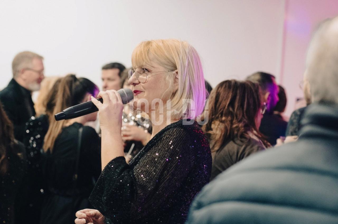 Femme blonde tenant un micro, parlant devant un groupe de personnes dans un espace intérieur éclairé.