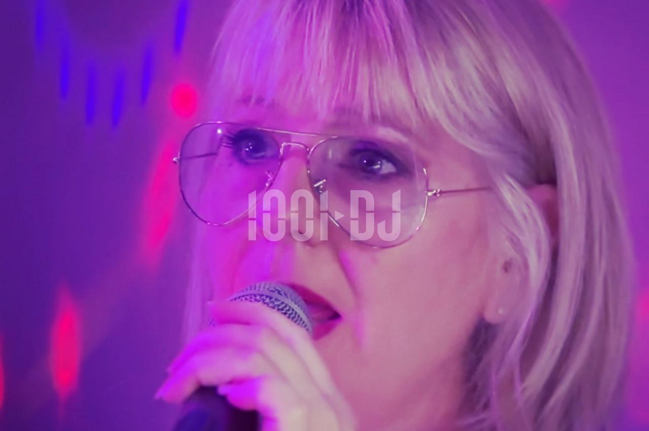 Femme blonde portant des lunettes, chantant au micro sous des lumières violettes et rouges.