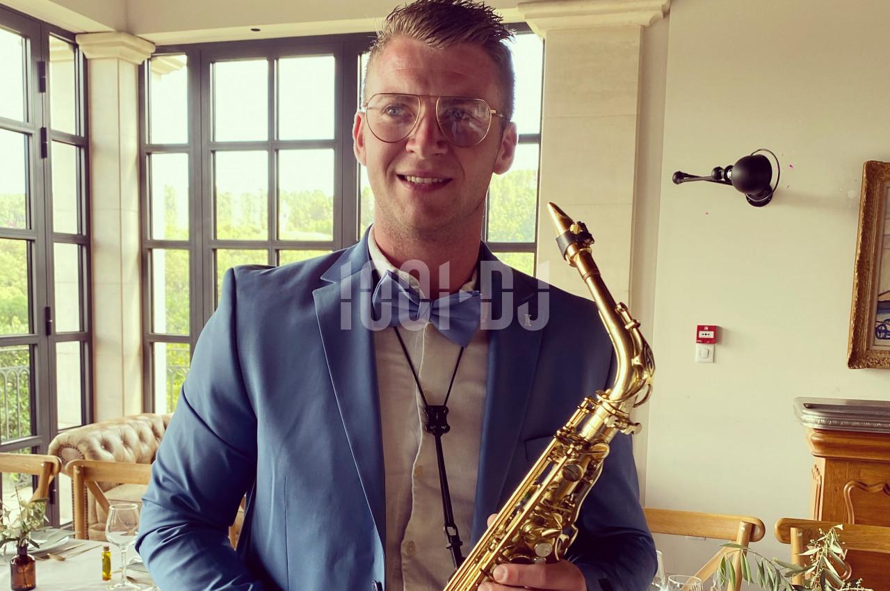 Un homme en costume bleu tient un saxophone dans un intérieur lumineux avec de grandes fenêtres.
