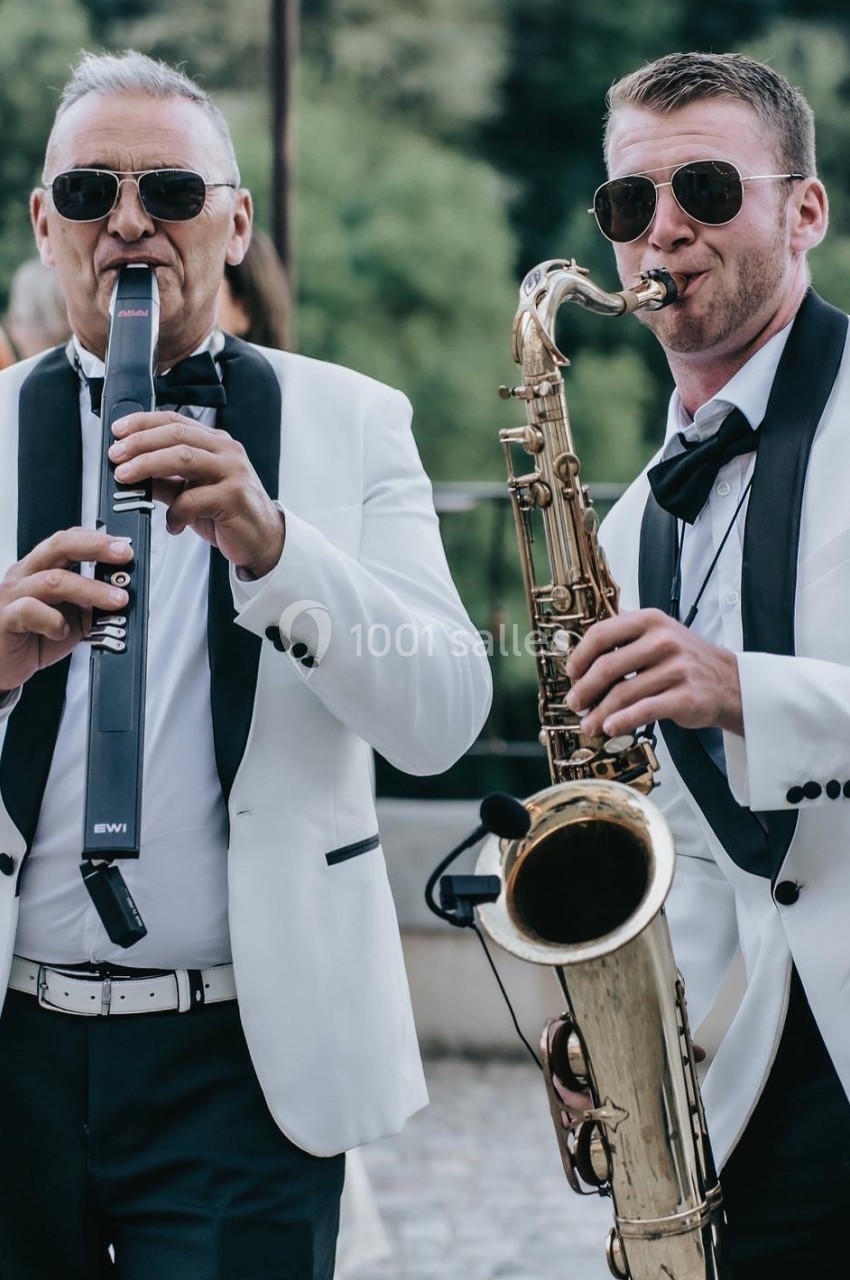Deux musiciens en costume blanc jouent du saxophone et d'une flûte électronique en extérieur.