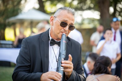 Un homme en costume bleu tient un saxophone sous une pergola décorée de fleurs blanches.