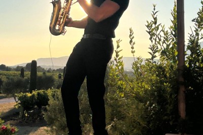 Un homme en costume bleu tient un saxophone sous une pergola décorée de fleurs blanches.