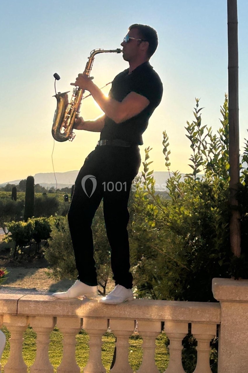 Un homme joue du saxophone debout sur une balustrade, avec un paysage ensoleillé en arrière-plan.