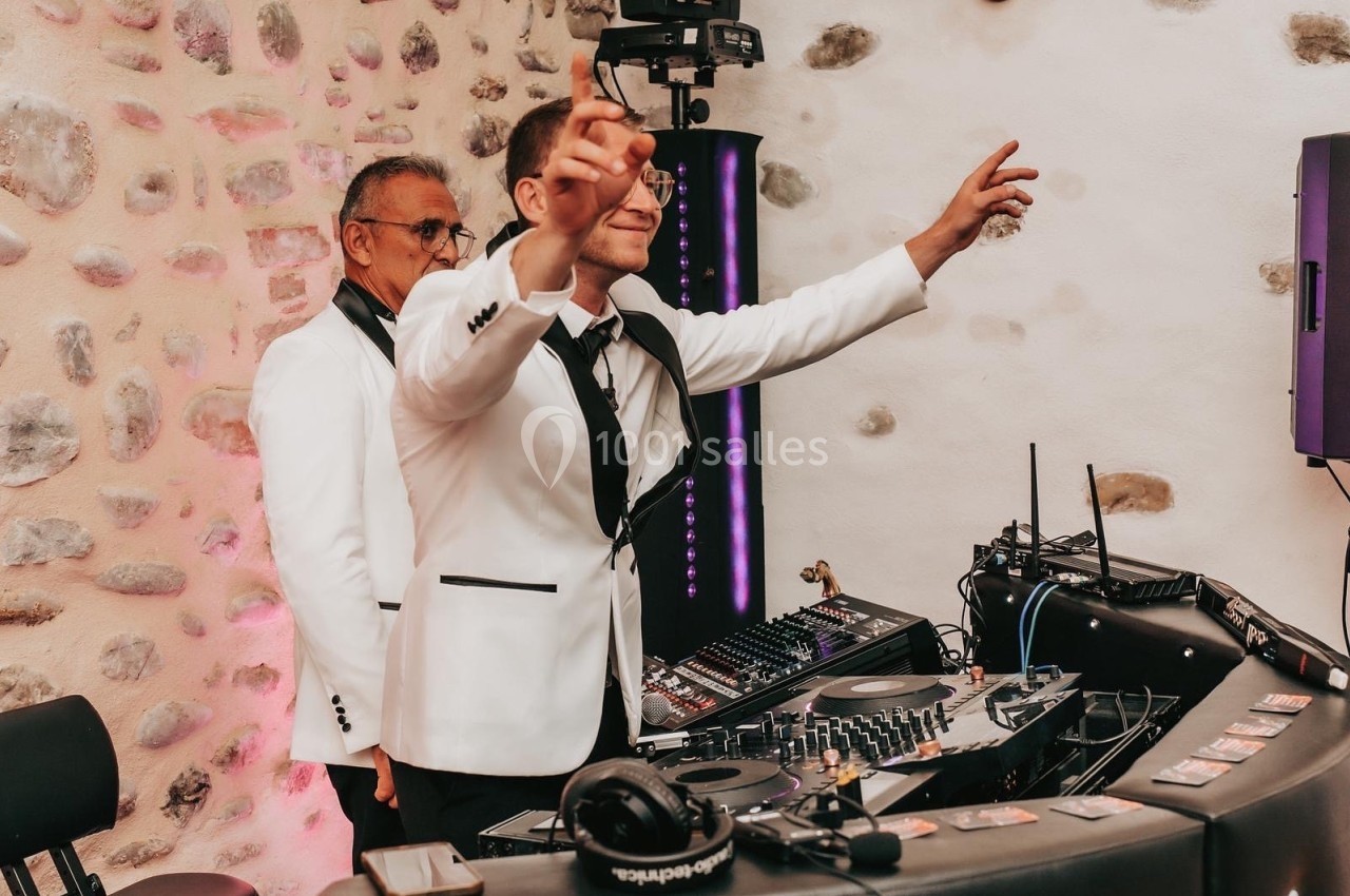 Deux DJ en costume blanc animent une soirée avec une table de mixage dans un décor en pierre apparente.