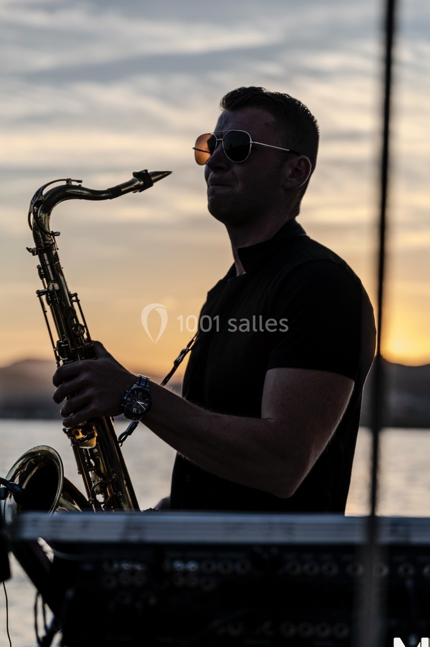 Un musicien joue du saxophone au coucher du soleil, avec un arrière-plan de ciel et d'eau.