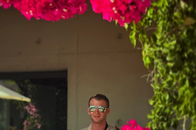 Un homme en costume bleu tient un saxophone sous une pergola décorée de fleurs blanches.