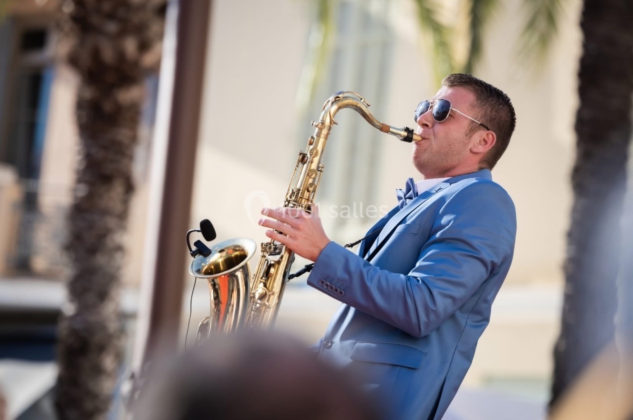 Un homme en costume bleu joue du saxophone en extérieur, sous un ciel ensoleillé, près de palmiers.