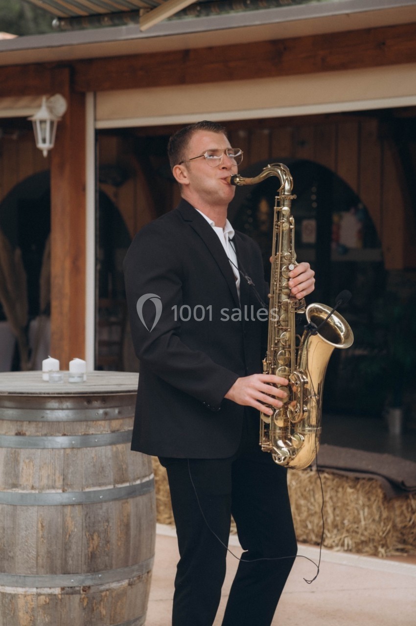 Un musicien en costume joue du saxophone devant un tonneau en bois, près d'un bâtiment en bois.