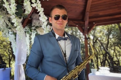Un homme en costume bleu tient un saxophone sous une pergola décorée de fleurs blanches.
