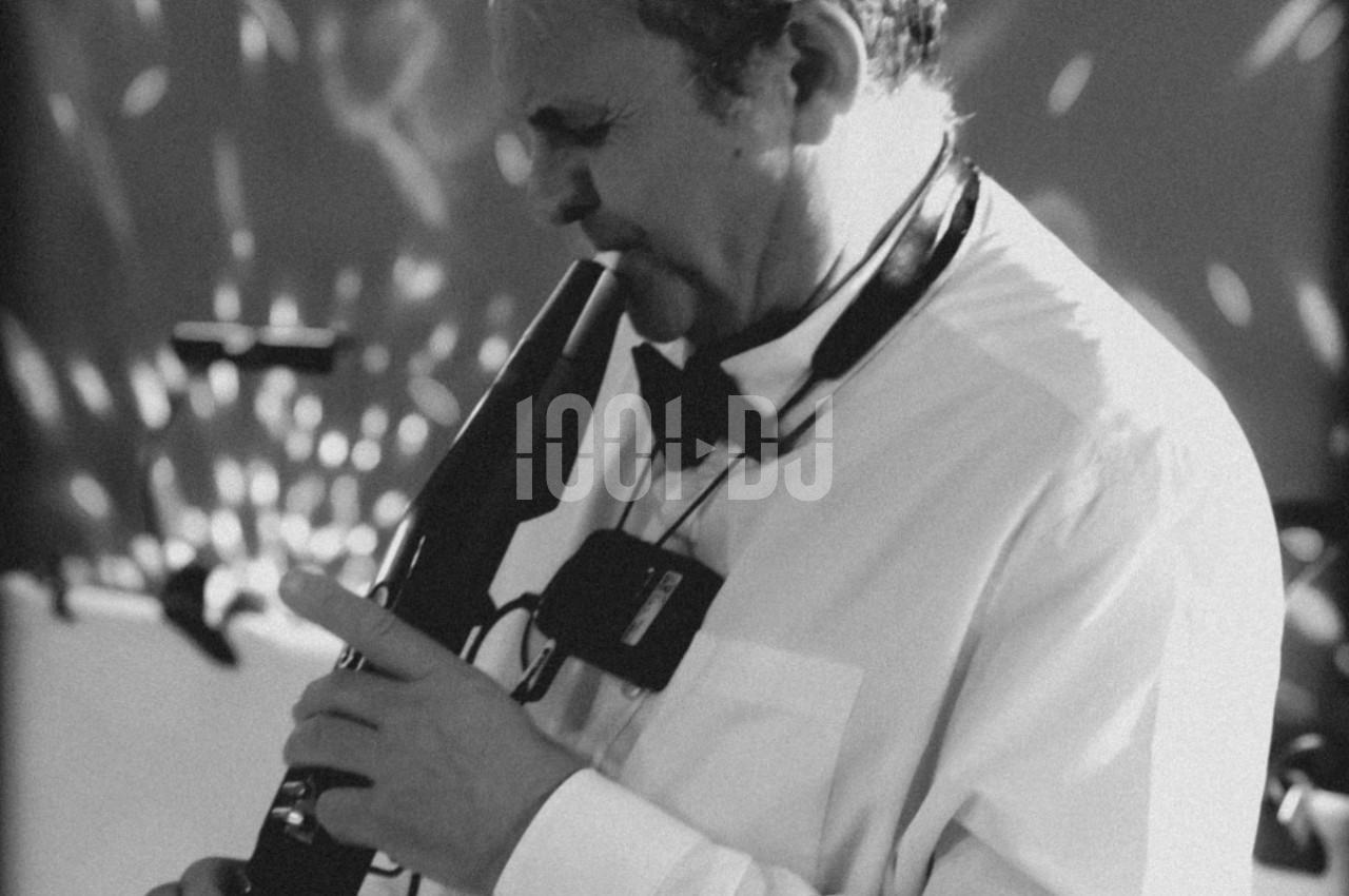 Un musicien en chemise blanche joue d'un instrument à vent électronique dans une ambiance lumineuse tamisée.