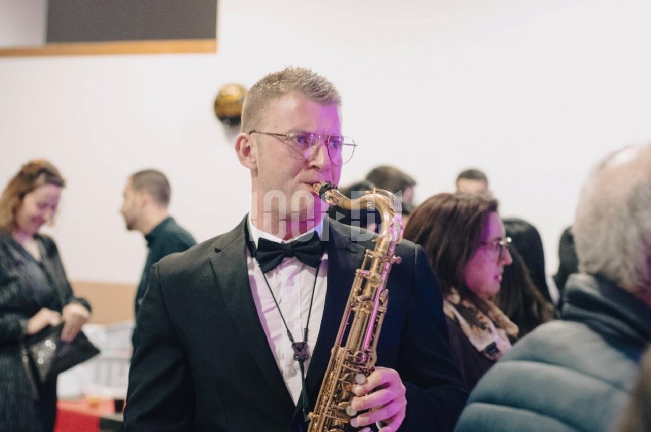 Un musicien en smoking joue du saxophone lors d'un événement avec des personnes en arrière-plan.