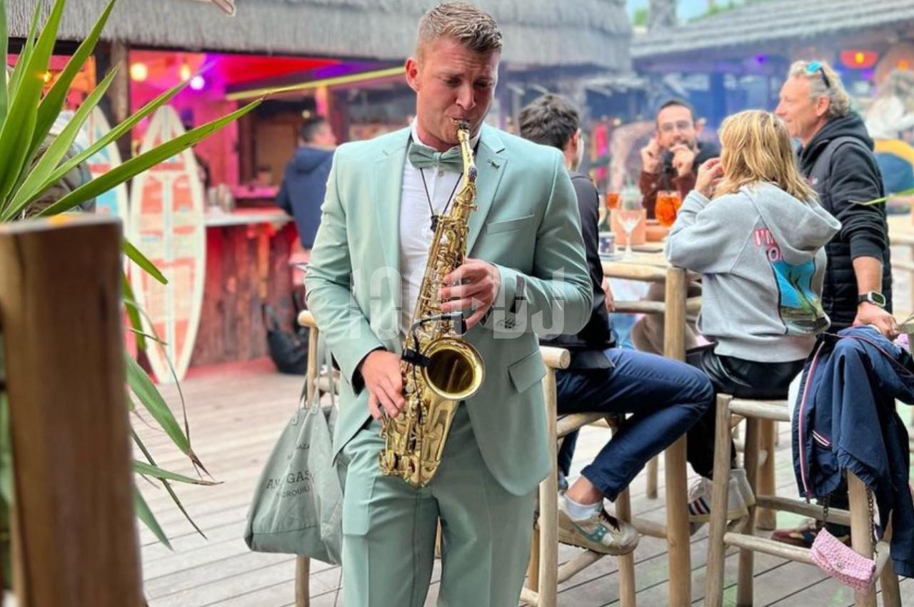 Un homme en costume vert joue du saxophone dans un bar en plein air, entouré de clients assis et debout.