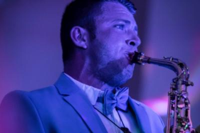 Un homme en costume bleu tient un saxophone dans un intérieur lumineux avec de grandes fenêtres.