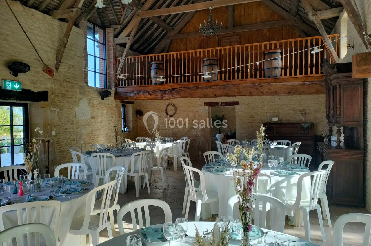 Salle de réception rustique avec tables dressées, chaises blanches, poutres apparentes et mezzanine en bois.