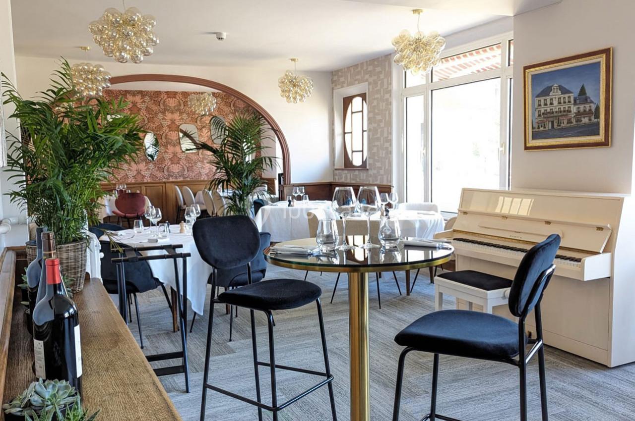 Salle de restaurant lumineuse avec tables dressées, piano blanc, plantes décoratives et grandes baies vitrées.