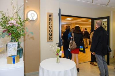Salle décorée avec des ballons roses, éclairage violet, deux personnes marchant au centre, tables hautes et chaises…