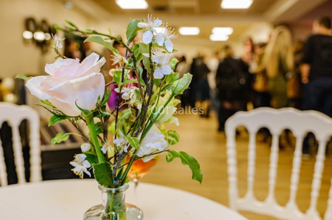 Un vase avec une rose rose et des fleurs blanches sur une table, avec des personnes floues en arrière-plan.