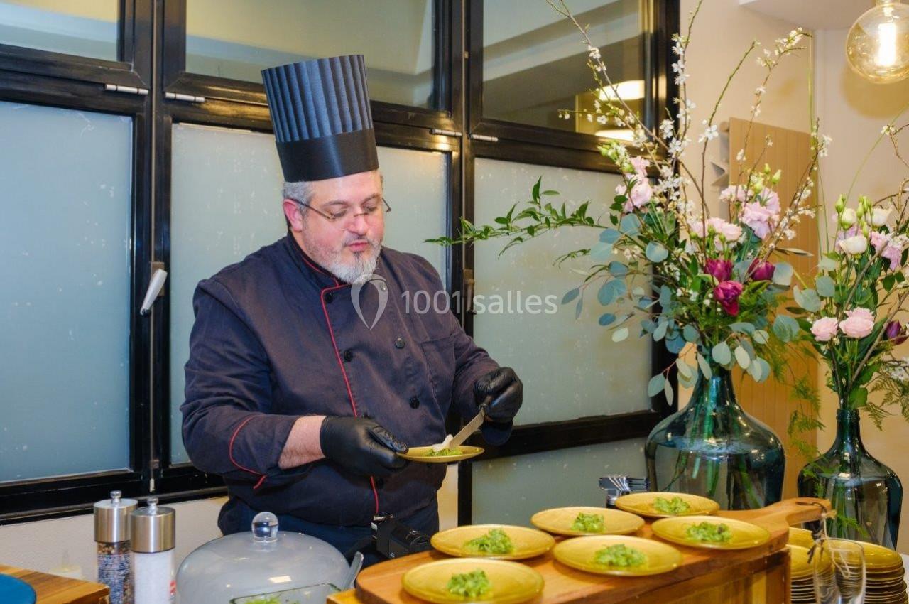 Un chef en tenue noire prépare des assiettes garnies de légumes verts dans une cuisine décorée de fleurs.