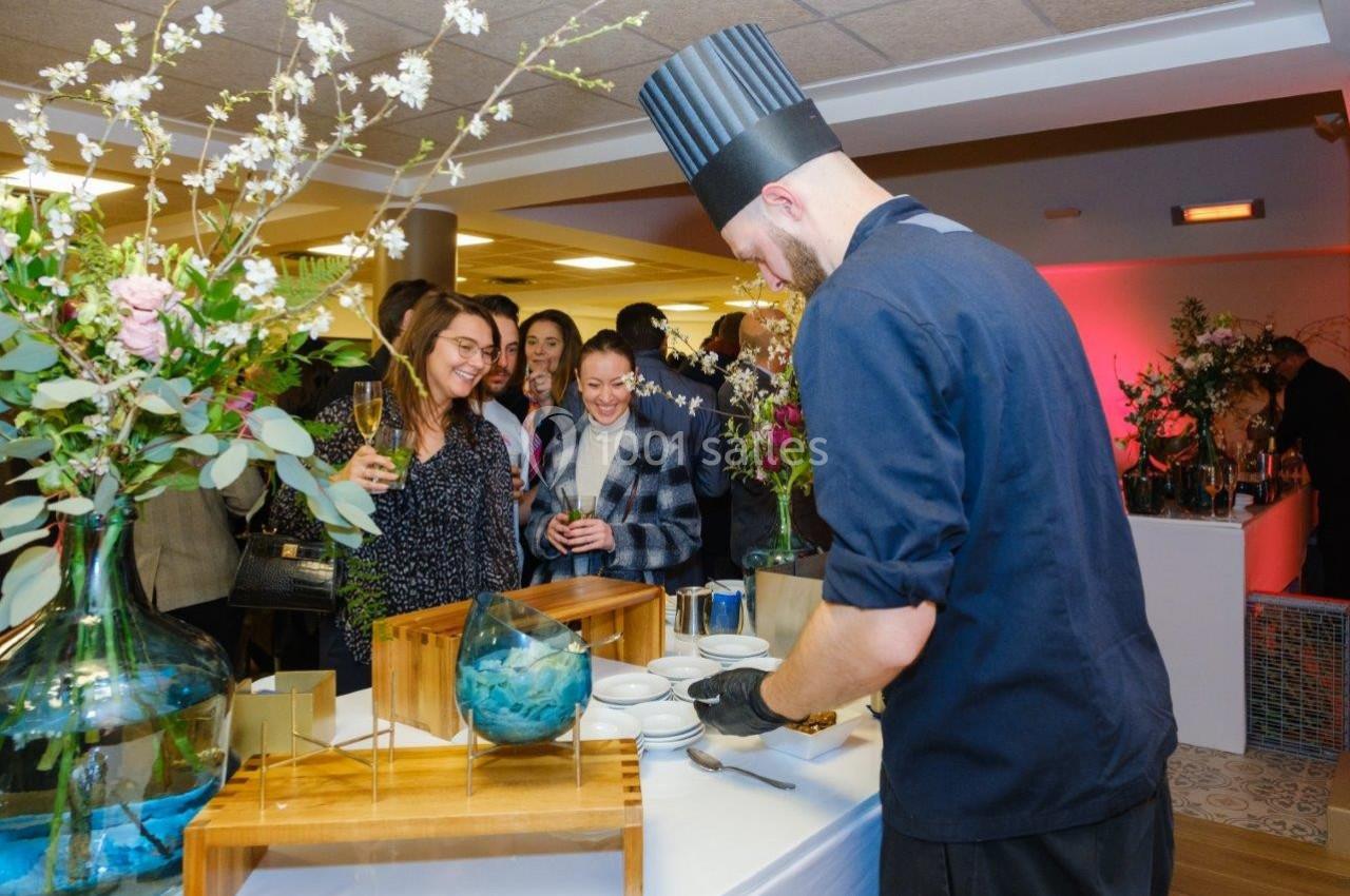 Un chef en tenue prépare un plat devant des invités souriants lors d'un événement avec une décoration florale.
