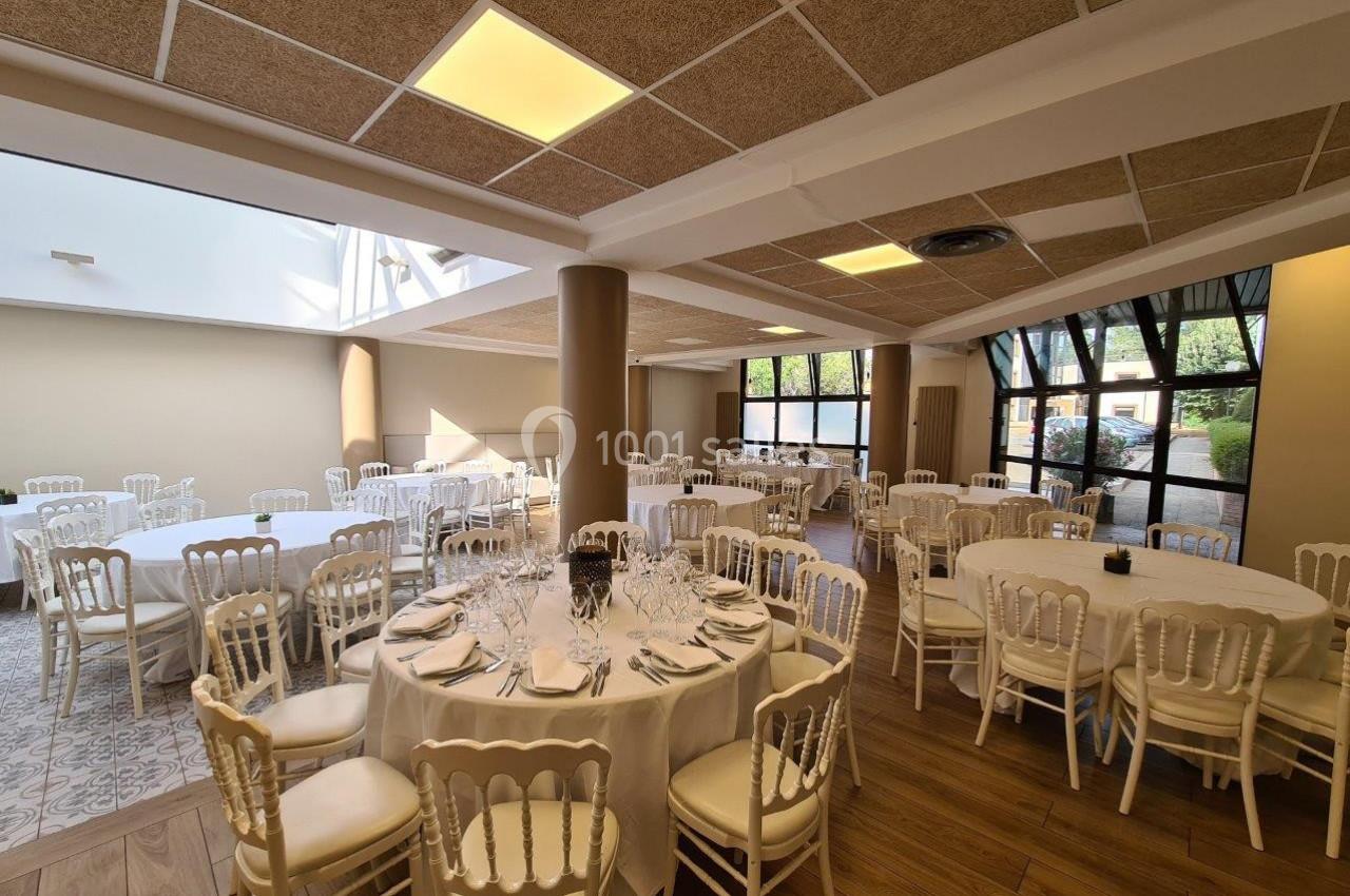 Salle de réception lumineuse avec tables rondes dressées, chaises blanches et grandes fenêtres donnant sur l'extérieur.