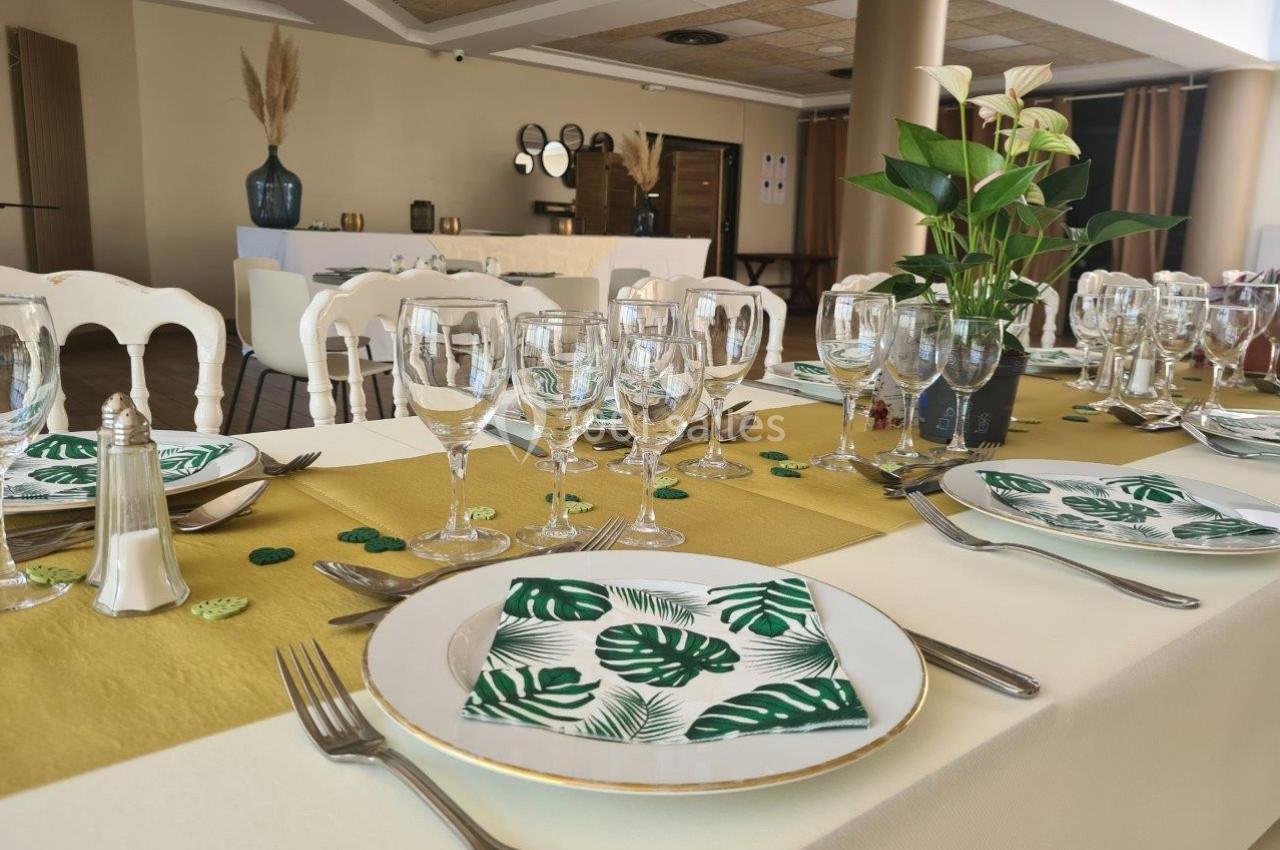 Table dressée avec nappes blanches, chemins de table dorés, vaisselle élégante et serviettes à motif de feuilles tropicales.