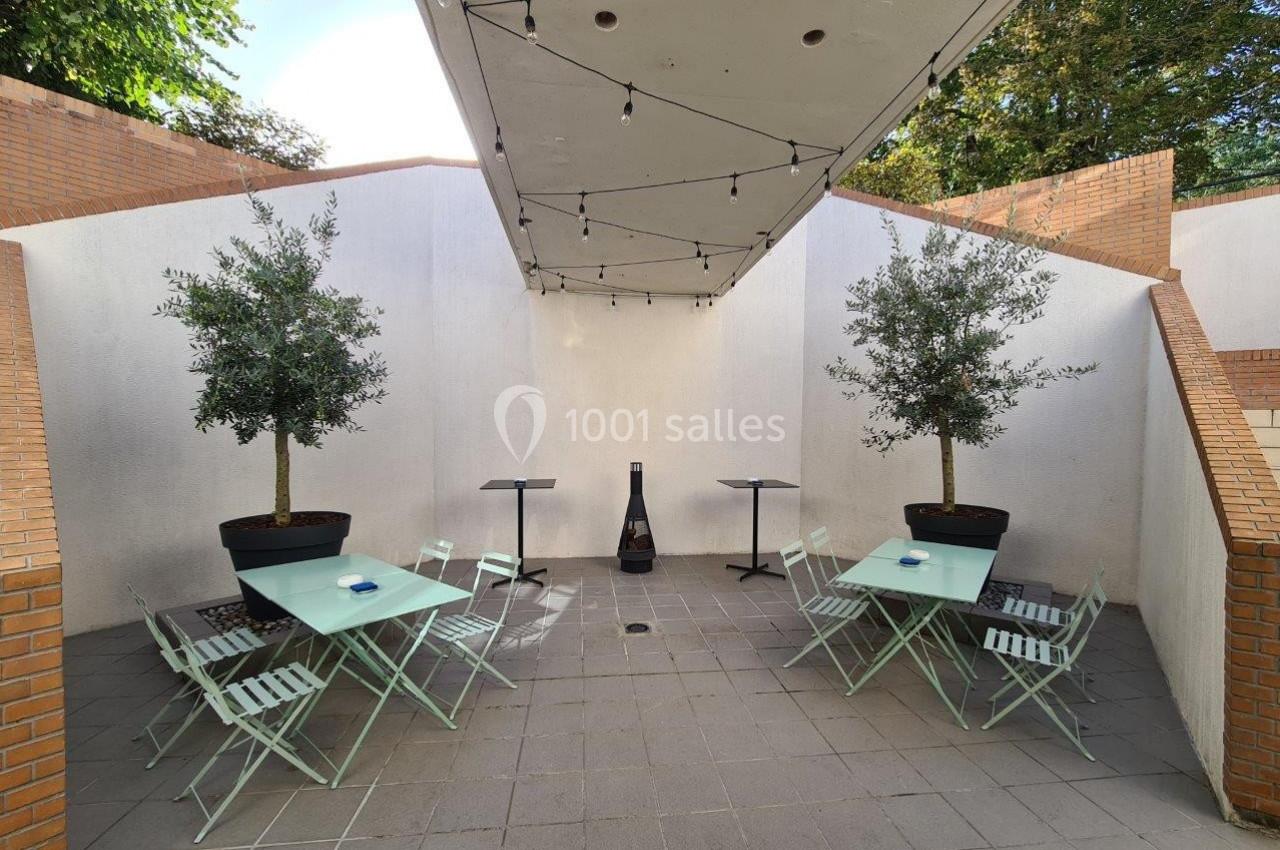 Terrasse extérieure avec tables, chaises pliantes vert clair, deux oliviers en pot et guirlandes lumineuses suspendues.