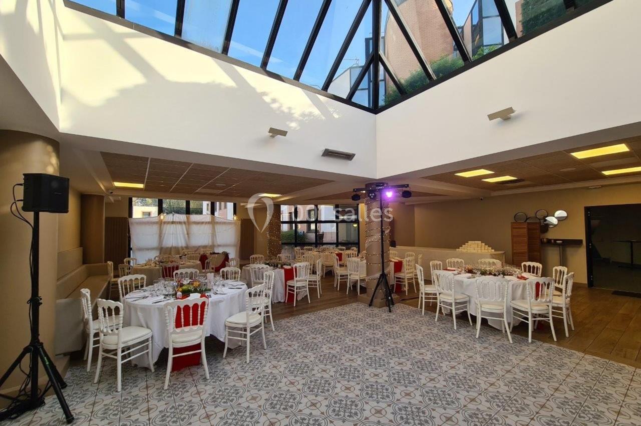Salle lumineuse avec verrière, tables rondes dressées avec nappes blanches et chaises, prête pour un événement.