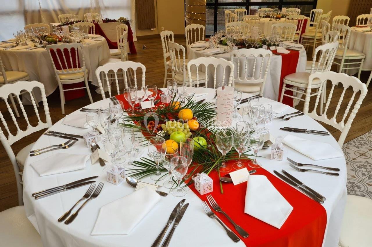 Table ronde décorée pour un événement, avec nappes blanches, chemin de table rouge, fruits et couverts élégamment disposés.