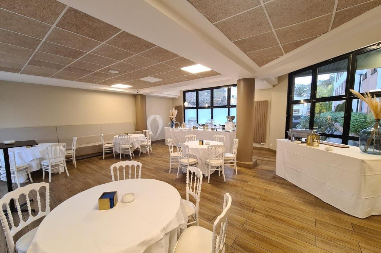 Salle lumineuse avec tables rondes dressées, chaises blanches et grandes fenêtres donnant sur l'extérieur.