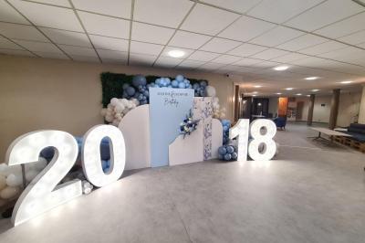 Salle décorée avec des ballons roses, éclairage violet, deux personnes marchant au centre, tables hautes et chaises…
