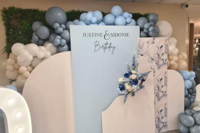 Salle décorée avec des ballons roses, éclairage violet, deux personnes marchant au centre, tables hautes et chaises…