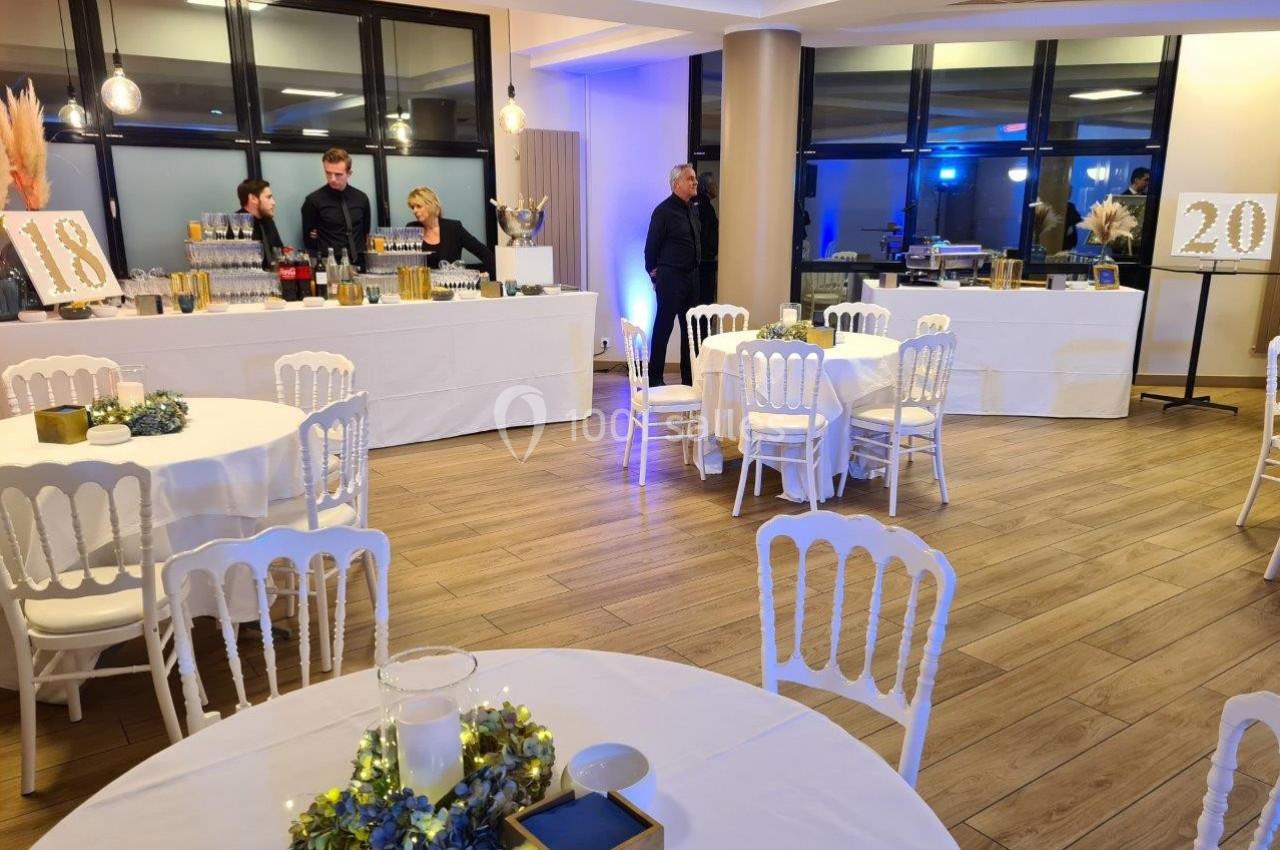 Salle décorée pour un événement avec tables rondes, chaises blanches et buffet en arrière-plan.