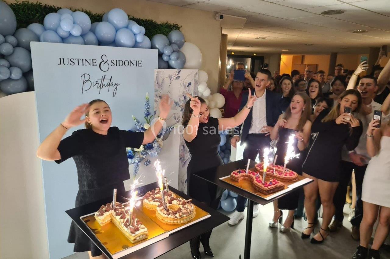 Deux femmes soufflent des bougies sur des gâteaux d'anniversaire, entourées d'invités dans une salle décorée.