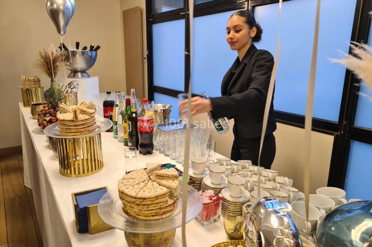Une femme en tenue professionnelle prépare des boissons sur une table garnie de snacks et de décorations festives.