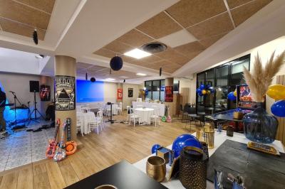 Salle décorée avec des ballons roses, éclairage violet, deux personnes marchant au centre, tables hautes et chaises…