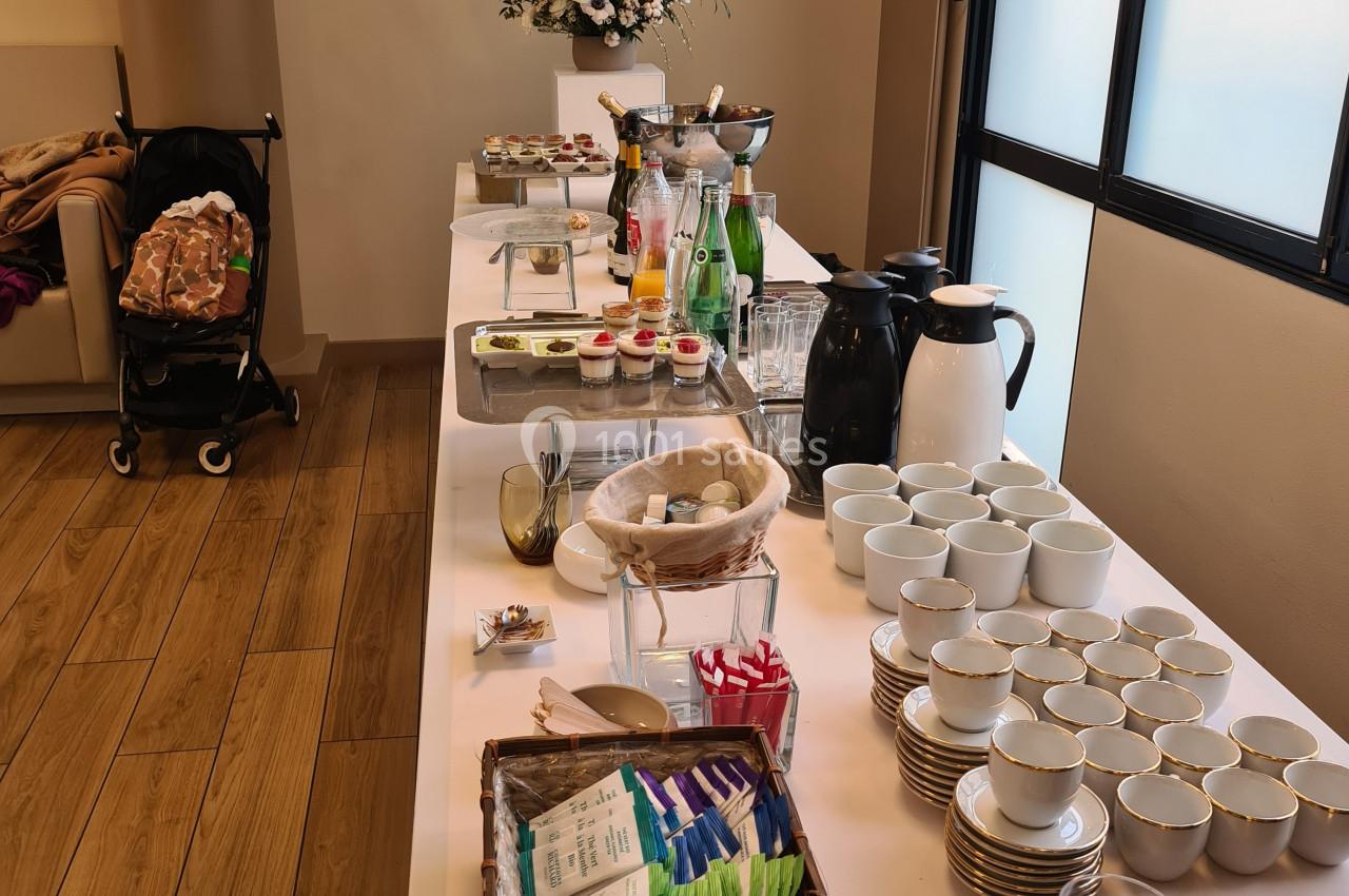 Table de buffet avec boissons, tasses, viennoiseries et fleurs, dans une salle lumineuse avec parquet et fenêtres.