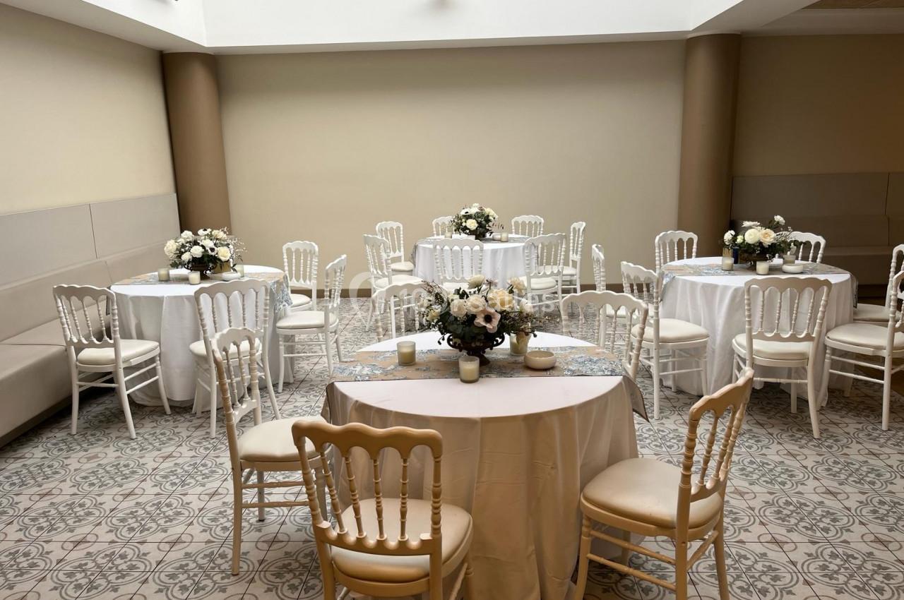 Salle lumineuse avec tables rondes décorées de nappes blanches et bouquets de fleurs, entourées de chaises élégantes.
