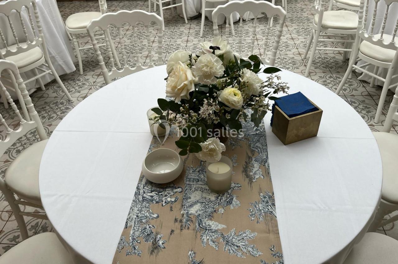 Table ronde décorée avec une nappe blanche, un chemin de table beige à motifs, des fleurs blanches et des bougies.