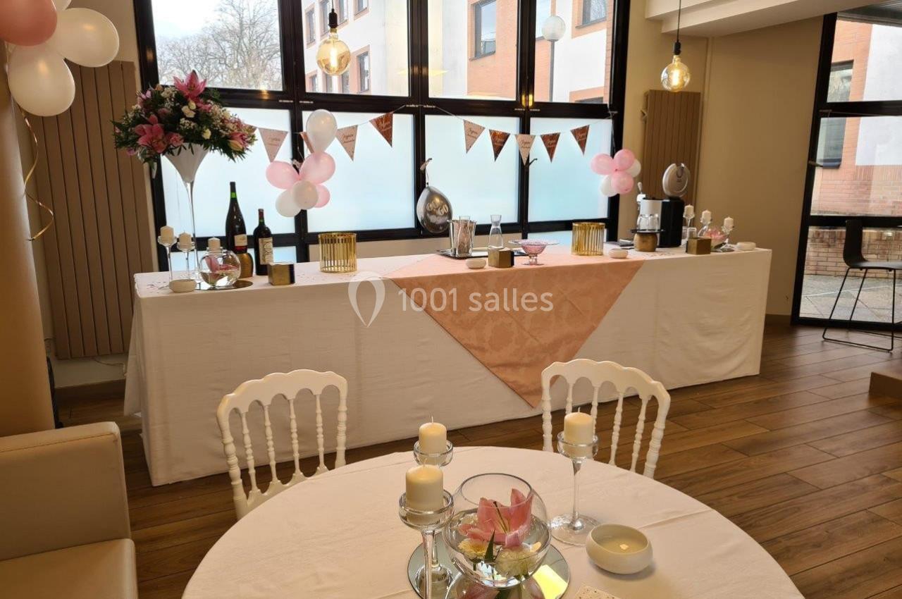 Table décorée pour un événement avec nappes blanches, fleurs, bougies et vaisselle, dans une salle lumineuse.