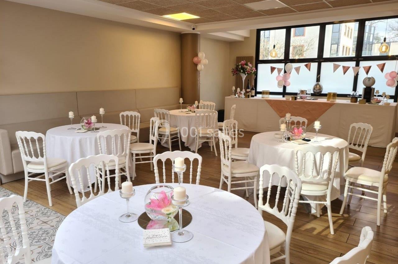 Salle décorée pour un événement, avec tables rondes, nappes blanches, chaises blanches et guirlandes en arrière-plan.