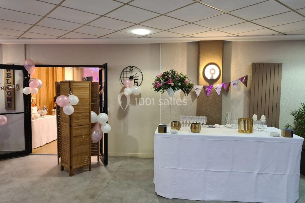 Salle décorée pour un événement avec table dressée, ballons, guirlandes et fleurs près d'une cloison en bois.