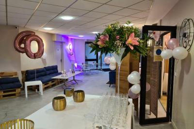 Salle décorée avec des ballons roses, éclairage violet, deux personnes marchant au centre, tables hautes et chaises…