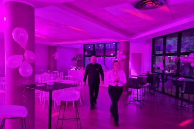 Salle décorée avec des ballons roses, éclairage violet, deux personnes marchant au centre, tables hautes et chaises…