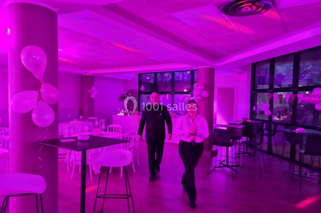 Salle décorée avec des ballons roses, éclairage violet, deux personnes marchant au centre, tables hautes et chaises…