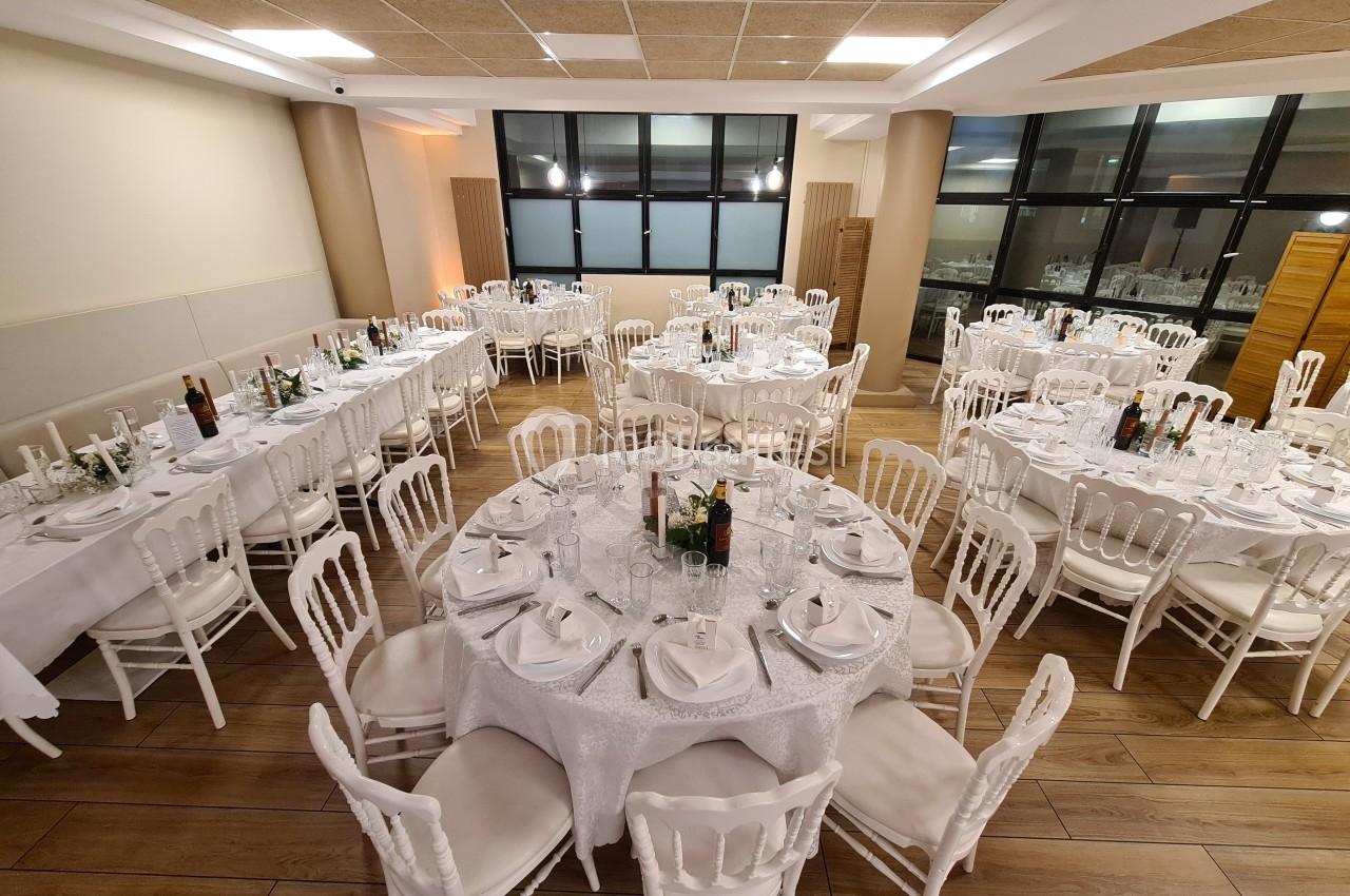 Salle de réception avec tables rondes et rectangulaires dressées, nappes blanches et chaises en bois clair.