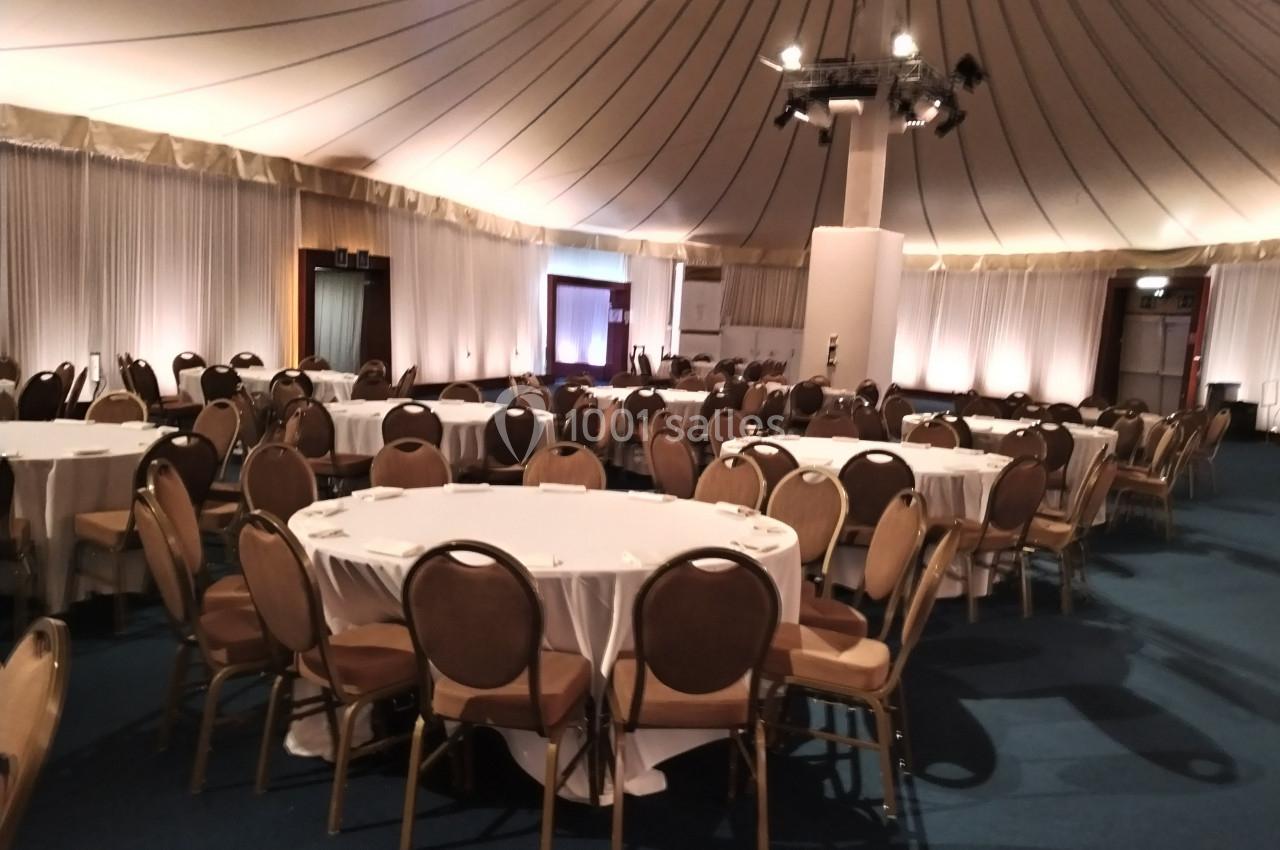 Salle de réception avec tables rondes dressées de nappes blanches et chaises disposées en cercle.