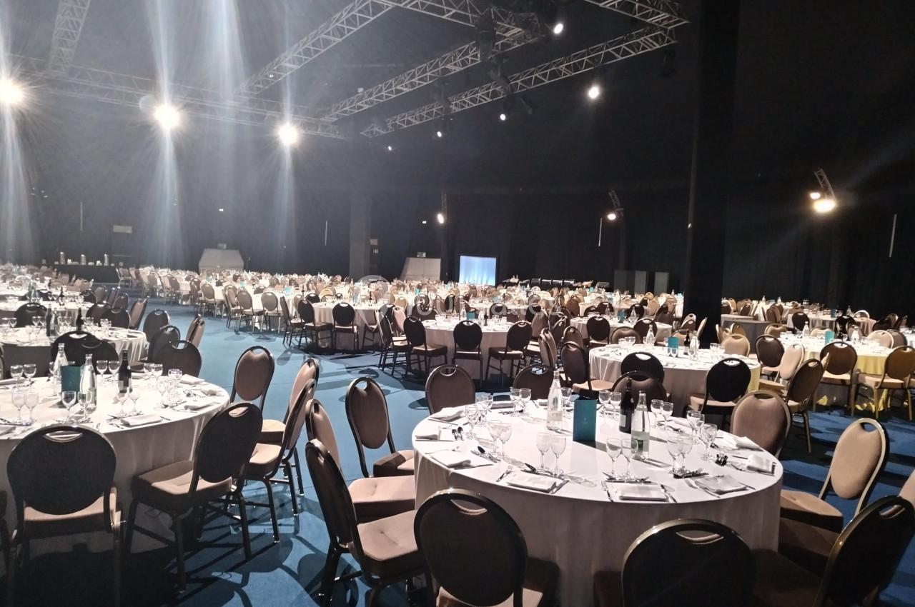 Salle de réception avec de nombreuses tables rondes dressées, éclairée par des projecteurs au plafond.